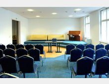 Ein kleiner Veranstaltungsraum mit blauen Stuhlreihen, einem erhöhten Podium mit einem Pult, zwei gelben Sofas und einem hölzernen Klavier im Hintergrund. Der Raum hat weiße Wände, mehrere Fenster auf der rechten Seite und eine moderne Deckenbeleuchtung.