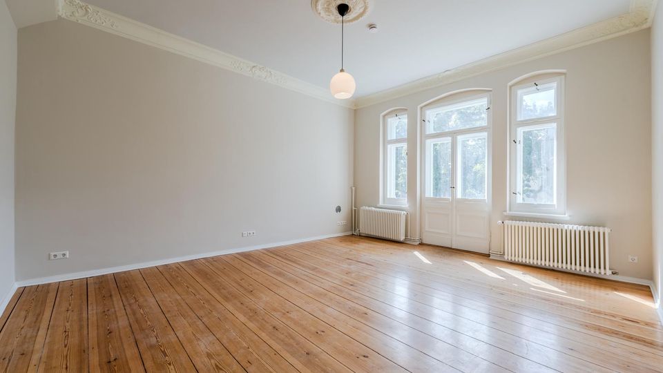 Heller, leerer Altbau-Raum mit breiten Holzdielen, hohen Decken mit Stuck und einer zentralen Pendelleuchte. An der Stirnseite befinden sich hohe, weiß gerahmte Fenster und eine verglaste Flügeltür, darunter weiße Heizkörper.