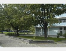 Ein moderner Wohnkomplex mit großen Fenstern und Balkonen, umgeben von einer begrünten Fläche mit großen, schattenspendenden Bäumen, gepflasterten Wegen und angelegten Grünflächen. Im Hintergrund sind geparkte Autos zu sehen.