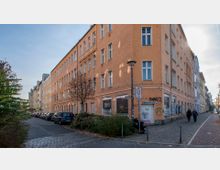 Eine Straßenecke in einem Wohnviertel mit einem vierstöckigen, orangefarbenen Mehrfamilienhaus, das Graffiti an unteren Fassadenbereichen zeigt. Entlang der Straße stehen parkende Autos, während der Gehweg rechts von zwei Personen genutzt wird; Straßenschilder markieren eine 30er-Zone.