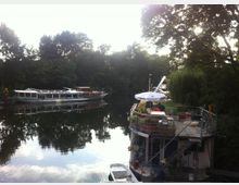 Hausboot mit einer Terrasse, auf der sich ein Sonnenschirm und Pflanzen befinden, liegt an einem Fluss vor einer bewaldeten Uferlandschaft; im Hintergrund ist ein weiteres, größeres Passagierboot zu sehen.