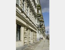 Straßenszene mit historischen Wohngebäuden, deren Fassaden mit Stuck und rechteckigen Fenstern verziert sind; Balkone sind mit Pflanzen und Blumen geschmückt. Der gepflasterte Gehweg führt entlang der Häuser, im Hintergrund sind Straßenlaternen und weitere Gebäude sichtbar.