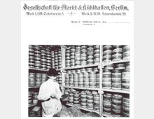 Historische Schwarz-Weiß-Aufnahme einer Lagerhalle mit hohen Regalen, die mit runden Metallbehältern gefüllt sind. Im Vordergrund steht ein Mann in weißem Kittel und Hut, der einen Behälter hält und mit einem Werkzeug darin arbeitet.