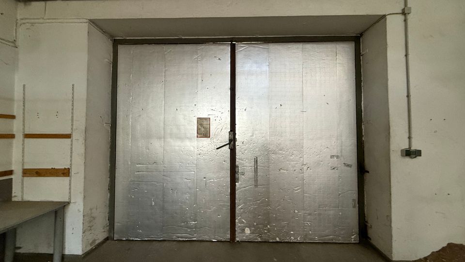 Innenansicht eines industriellen Lager- oder Garagenraums mit großer zweiflügeliger Metalltür in einer Wandnische. Sichtbare Deckenrohre und Kabelkanäle sowie schlichte weiße Wände und Betonboden prägen den Raum.