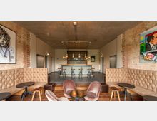 Ein modern gestalteter Lounge-Bereich mit einer Bar im Hintergrund, umgeben von freiliegenden Ziegelwänden und Betondecke. Gepolsterte Sitzbänke und Sessel mit kleinen runden Tischen stehen um die Bar, die von Hockern und hängenden Lampen eingerahmt ist; Wände sind mit Kunstwerken dekoriert.