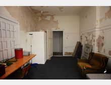 Ein einfacher Raum mit abgeblätterter Wandfarbe und abgenutztem Zustand. Im Raum befinden sich ein Holztisch mit Gegenständen, ein alter Sessel, ein großer Schrank und ein Schild, während der Boden mit Teppich bedeckt ist.