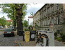 Straßenansicht mit historischen Wohnhäusern auf der rechten Seite, einer Baumreihe entlang des Gehwegs und geparkten Autos auf der linken Seite. Im Vordergrund stehen Mülltonnen sowie ein Stromkasten mit Graffiti, und im Hintergrund sind weitere Gebäude und eine Tankstelle zu erkennen.