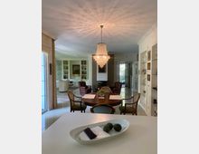 Wohnzimmer und Essbereich in einem offenen, hellen Raum mit glänzendem Boden. Im Vordergrund ein Esstisch aus Holz mit Stühlen und einer Hängelampe aus Kristall; im Hintergrund sind ein Sitzbereich mit Sesseln, ein Bücherregal und ein Fenster mit Blick ins Freie sichtbar.