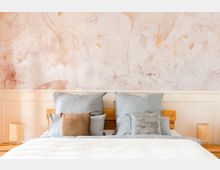 Schlafzimmer mit einem hölzernen Bettgestell, blaugrauer Bettwäsche und beigen sowie gemusterten Zierkissen. Im Hintergrund eine Wand mit dezentem floralen Muster in Pastelltönen, flankiert von zwei Nachttischen mit geflochtenen Lampenschirmen.