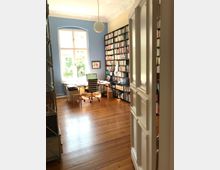 Ein heller, gemütlicher Arbeitsraum mit Holzboden, einem großen Fenster mit weißem Rahmen, einer blauen Wand und hohen Bücherregalen voller Bücher. In der Mitte steht ein Schreibtisch mit Laptop und Drehstuhl, über dem eine runde Hängelampe angebracht ist.