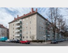 Eckansicht eines mehrstöckigen Wohnhauses aus der Nachkriegszeit mit heller Fassade und roten Ziegeldächern. Das Gebäude hat Balkone mit grünen Brüstungen, vor denen parkende Autos sowie kahler Baumbestand und eine Gehwegkreuzung zu sehen sind.