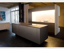 Moderne Küche in einem Loft mit freistehender Kücheninsel, eingelassenem Kochfeld und indirekter Beleuchtung. Im Hintergrund sind Backsteinwände, eine große Fensterfront und ein Teil eines Wohnbereichs erkennbar.