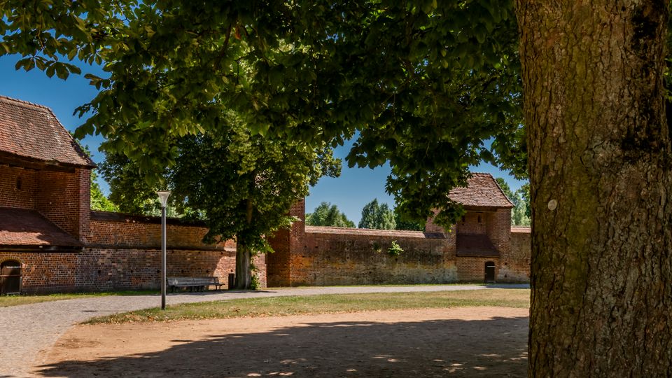 Blick auf eine lange historische Backstein-Stadtmauer mit zwei turmartigen Torbauten und kleinen Durchgängen, davor ein geschwungener Kiesweg und eine Sitzbank. Große Bäume rahmen die Szene und spenden Schatten über einer offenen Grün- und Sandfläche.