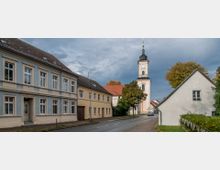 Straßenszene in einer Kleinstadt mit historischen Gebäuden, darunter ein hellblaues und ein beigefarbenes Haus auf der linken Seite. Im Hintergrund steht eine Kirche mit einem hohen Turm und einer Uhr, umgeben von Bäumen und einem gepflasterten Gehweg entlang der Straße.
