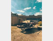 Industriehof mit Schotterboden, umgeben von Containern und Lagerhallen, teils mit Graffiti. Im Vordergrund steht ein ausgebranntes Autowrack in einem Metallrahmen neben Holzpaletten unter blauem Himmel mit Wolken.