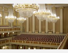 Innenansicht eines historischen Konzertsaals mit weißen Säulen, reich verzierten goldenen Elementen und roten Polstersitzen. Mehrere große, prunkvolle Kristalllüster hängen von der Decke, während Vorhänge die hohen Wände bedecken.