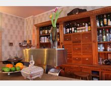 Ein gemütlicher Barbereich mit einer hölzernen Rückwand, die aus Regalen mit Flaschen und kleinen Schubladen besteht. Im Vordergrund steht eine silberne Kaffeemaschine mit einer Vase mit frischen Blumen, daneben eine Schale mit Zitrusfrüchten.