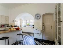 Helle Küche mit weißer Einrichtung, einem schwarz-weiß karierten Boden und einem großen Durchgangsbogen. Links befinden sich Schränke und eine Arbeitsfläche mit Obst und zwei Barhockern, rechts ein kleines Waschbecken und eine Tür, im Hintergrund ein Fenster mit Sprossen und Pflanzen.