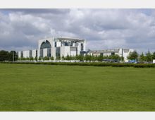 Modernes Regierungsgebäude mit einer markanten weißen Fassade, großen Glasflächen und einem halbkreisförmigen Oberteil, umgeben von einer Grünfläche und jungen Bäumen. Der Himmel ist teils bewölkt, und im Hintergrund sind Flaggen und einige Fahrzeuge sichtbar.