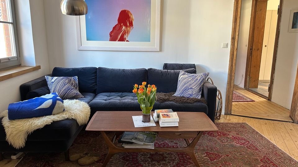 Gemütliches Wohnzimmer mit dunkler Eckcouch, Kissen und Decken sowie einem niedrigen Holzcouchtisch mit Tulpen und Büchern auf einem gemusterten Teppich. An der weißen Wand hängt ein großes gerahmtes Bild; links fällt Tageslicht durch ein Fenster, rechts führt ein rustikaler Holztürrahmen in einen Flur mit Holzdielen.