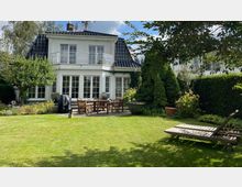 Zweistöckiges weißes Einfamilienhaus mit schwarzem Ziegeldach und großen bodentiefen Fenstern, umgeben von einem gepflegten Garten mit Rasen, Bäumen und Sträuchern. Im Vordergrund stehen Holzmöbel auf einer Terrasse sowie zwei Liegestühle auf dem Rasen.