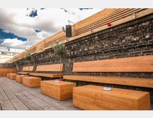 Sitzbereich im Außenbereich mit Holzbänken und -tischen vor einer Ziegelmauer; kleine Olivenbäume sind entlang der Sitze aufgereiht, und darüber verläuft eine Holzverkleidung. Der Boden besteht aus Holzdielen, und der Himmel ist teilweise bewölkt.