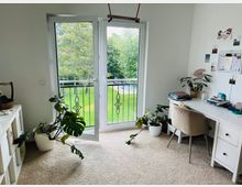 Helles Arbeitszimmer mit weißer Einrichtung, bestehend aus einem Schreibtisch und einem Regal. Eine Glastür mit Blick auf einen grünen Garten führt auf einen kleinen Balkon mit schmiedeeisernem Geländer, im Raum stehen mehrere Zimmerpflanzen.