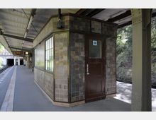 Bahnhof mit überdachtem Bahnsteig, einem Kiosk oder Betriebsraum mit brauner Holztür und gekachelter Fassade; im Hintergrund Gleise, ein Tunnel und eine Anzeigetafel.