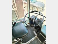 Innenraum eines Fahrerkabinenbereichs in einem Bus oder Lastwagen mit einem schwarzen, gepolsterten Sitz, einem großen Lenkrad und einem einfach gestalteten Armaturenbrett mit Tachometern. Im Hintergrund sind durch die Frontscheibe ein geparktes Auto und ein Teil des Außenbereichs sichtbar.