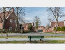 Straßenszene in einer Wohngegend mit Reihen von Backsteinhäusern im traditionellen Stil, roten Ziegeldächern und weißen Fensterrahmen. Im Vordergrund befindet sich eine grüne Parkbank an einem Gehweg, umgeben von jungen Bäumen und Grünflächen.