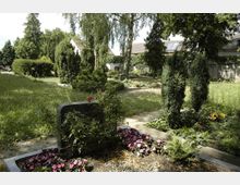 Ein gepflegter Friedhof mit mehreren Grabsteinen, umgeben von Blumenbeeten, kleinen Büschen und hohen Bäumen. Im Hintergrund sind Gebäude mit Ziegeldächern und eine Hecke zu sehen.