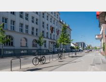 Straßenansicht mit einem weißen, mehrstöckigen Gebäude, das als Rathaus dient. Vor dem Gebäude befinden sich Bäume, Fahrradständer mit geparkten Fahrrädern, sowie eine saubere Gehweg- und Straßenfläche, im Hintergrund sind weitere Gebäude sichtbar.