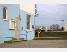 Ein einfacher, hellblauer Gebäudezugang mit Treppe und blauem Geländer, daneben ein Schaukasten und ein "Halteverbot"-Schild. Im Hintergrund sind eine Außentreppe mit weißem Geländer, Bäume und ein Lautsprecher auf einem Mast zu sehen.