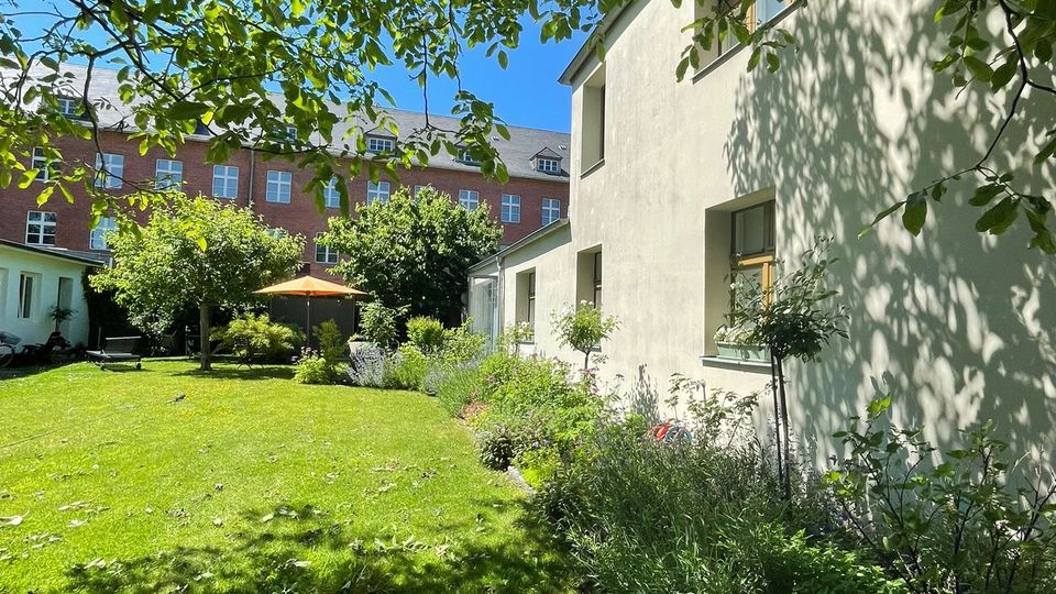 Innenhofgarten mit großer Rasenfläche, umgeben von Bäumen und Sträuchern, mit einer Terrasse und Sonnenschirm im Hintergrund. Rechts steht ein hell verputztes Wohngebäude mit mehreren Fenstern, im Hintergrund ist ein mehrstöckiges Backsteingebäude zu sehen.