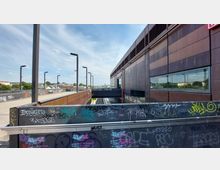 Außenansicht eines modernen Bahnsteigbereichs mit einem flachen Gebäude aus dunklen Metallpaneelen, einer verglasten Brücke darüber und Graffiti an Geländern. Im Hintergrund sind mehrere Straßenlaternen und teilweise bewachsene Dachflächen zu sehen.