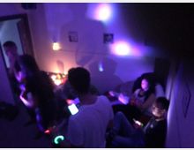 Mehrere Personen befinden sich in einem abgedunkelten Raum mit bunten Lichteffekten an den Wänden. Einige sitzen auf Möbeln und nutzen ihre Smartphones, während andere stehen oder sich bewegen.