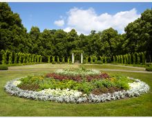 Runder Parkbereich mit bunt bepflanztem Blumenbeet im Vordergrund, umgeben von Rasenflächen und einem symmetrischen Laubengang aus überwucherten Bögen. Im Hintergrund stehen weiße Säulen unter einer Pergola vor einer dicht bewaldeten Kulisse.