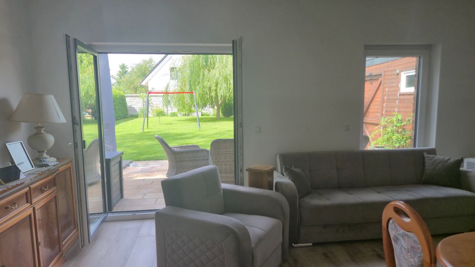 Helles Wohnzimmer mit grauem Sofa und Sessel sowie Holzkommode; eine große, geöffnete Terrassentür führt auf eine gepflasterte Terrasse und in einen grünen Garten. Draußen sind Rattanmöbel, ein Rasen mit Schaukelgestell und Bäume zu sehen.