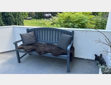 Eine graue Holzbank mit zwei dunklen Kissen und einem braunen Fell darauf steht auf einer gepflasterten Terrasse. Im Hintergrund ist ein Garten mit Rasen, Büschen und Bäumen zu sehen.