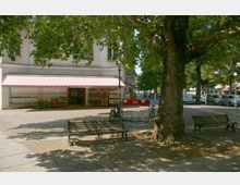 Ein kleiner Platz mit Kopfsteinpflaster, umgeben von Bäumen. Im Vordergrund stehen Holzbänke und ein großer Baum, im Hintergrund befindet sich ein Geschäft mit rot-weiß gestreifter Markise.