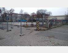 Eingezäuntes, brachliegendes Gelände mit Pflastersteinen im Vordergrund, Betonbarrieren und Metallzäunen, sowie schwarz-gelb markierten Absperrpfeilern. Im Hintergrund sind mehrere Bäume und Gebäude, teils mit Graffiti, sowie blauer Container zu sehen.