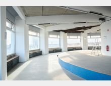 Offener, moderner Innenraum mit großen Fenstern, die einen Blick auf eine Stadtlandschaft ermöglichen. Der Raum ist hell, hat helle Wände, eine abgerundete, erhöhte Plattform im Vordergrund und eine Leiter daneben; die Decke zeigt sichtbare Betonstrukturen.