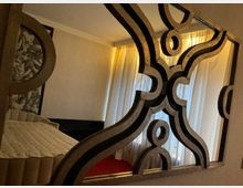 Spiegel mit dekorativen, geschwungenen Rahmen, der einen Teil eines Schlafzimmerinterieurs reflektiert. Im Hintergrund sind Vorhänge, ein Bett mit heller Bettdecke und eine Wand mit abstraktem Muster zu sehen.