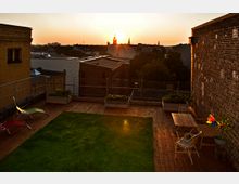 Dachterrasse mit einer Holzbodenfläche, einem kleinen Rasenstück, Sitzmöbeln und Pflanzkästen, umgeben von Backsteinmauern; im Hintergrund Blick auf eine Stadtlandschaft mit Silhouetten von Gebäuden und Kirchtürmen bei Sonnenuntergang.