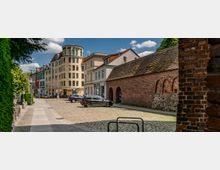 Straßenszene in einer Altstadt mit Kopfsteinpflaster, links ein moderneres Gebäude mit der Aufschrift „Haus am Steintor“ und rechts eine alte Ziegelmauer mit Rundbögen. Im Hintergrund stehen weitere klassische Stadtgebäude, und einige Autos sind auf dem Platz geparkt.