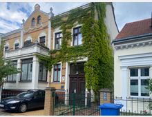 Wohnhaus mit gelber Klinkerfassade, dekorativen weißen Elementen und überwachsenem Efeu an der rechten Gebäudeseite. Vor dem Haus stehen ein geparkter schwarzer PKW und eine blaue Mülltonne, umgeben von einem Metallzaun.