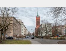 Straßenszene in einer Wohngegend mit mehrstöckigen Wohnhäusern und einem roten Kirchturm mit grünem Spitzdach im Hintergrund. Im Vordergrund befinden sich ein Gehweg, parkende Autos, Bäume ohne Laub und ein kleiner Spielplatz.