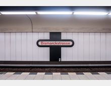 U-Bahn-Station mit einem weißen, vertikal gerillten Wandpaneel, in der Mitte ein schwarzes Kreuz mit dem Stationsnamen „Bismarckstraße“ in roter Schrift auf weißem Hintergrund. Darunter befinden sich Gleise und ein Bahnsteig mit schwarz-weiß gemusterten Fliesen.