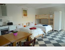 Ein einfacher Schlafraum mit schwarz-weiß kariertem Boden, ausgestattet mit einem Etagenbett, zwei Einzelbetten, einem Esstisch mit Stühlen und einer kleinen Küchenzeile mit Herd und Kühlschrank. An der Wand hängt ein farbiges Bild, und ein weißer Schrank steht in der Ecke des Raumes.