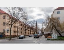 Straßenszene in einem Wohngebiet mit mehrstöckigen beige-gelben Wohnhäusern auf beiden Seiten, teilweise verdeckt durch unbelaubte Bäume. Im Hintergrund ragt der spitze Kirchturm einer Kirche auf, während parkende Autos entlang der Straße und ein Gehweg sichtbar sind.
